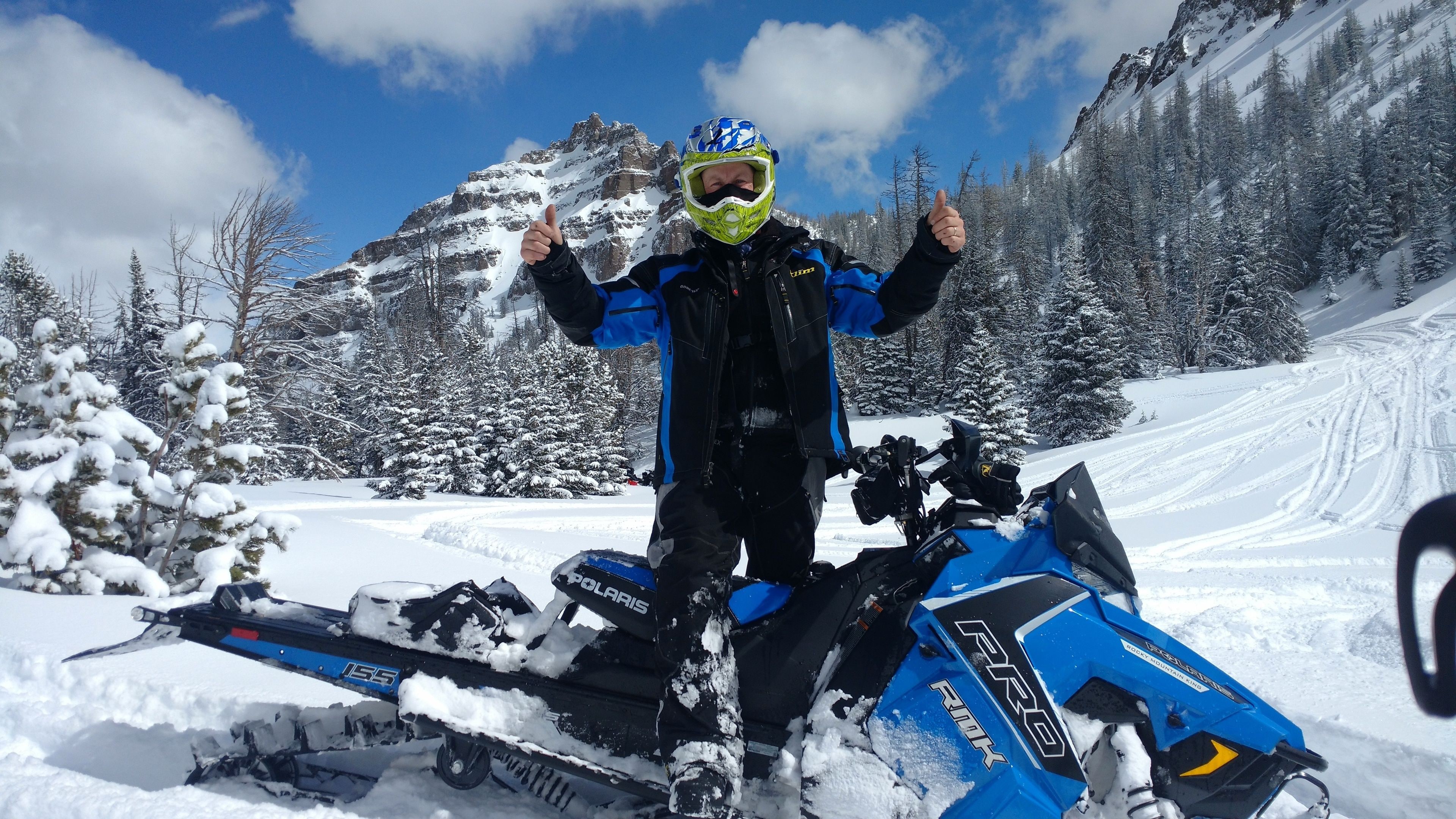 Una persona de pie junto a un vehículo para nieve en un paisaje nevado, con montañas al fondo.