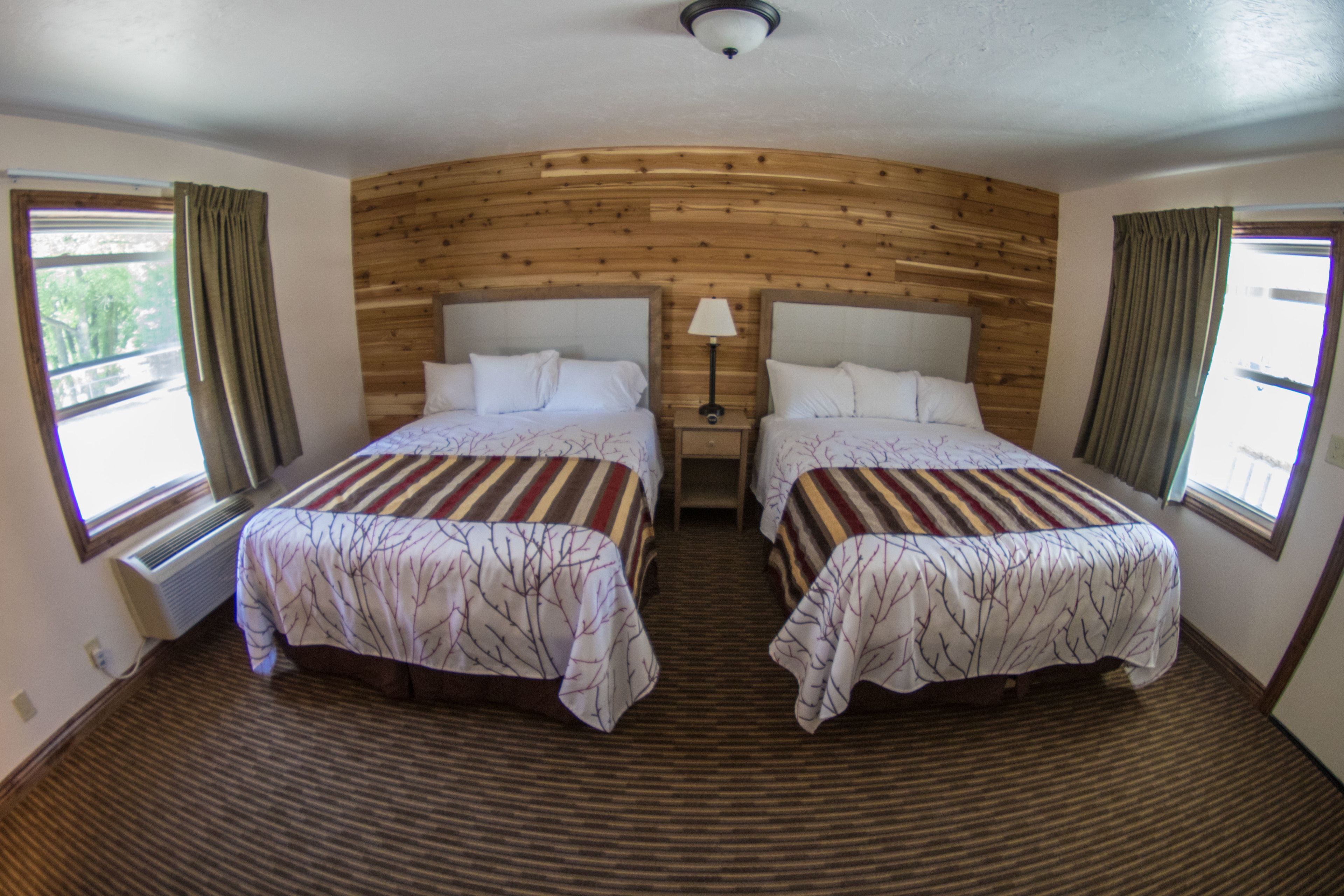 A room with two beds, wooden paneling on the walls, a window with curtains, and a nightstand with a lamp.