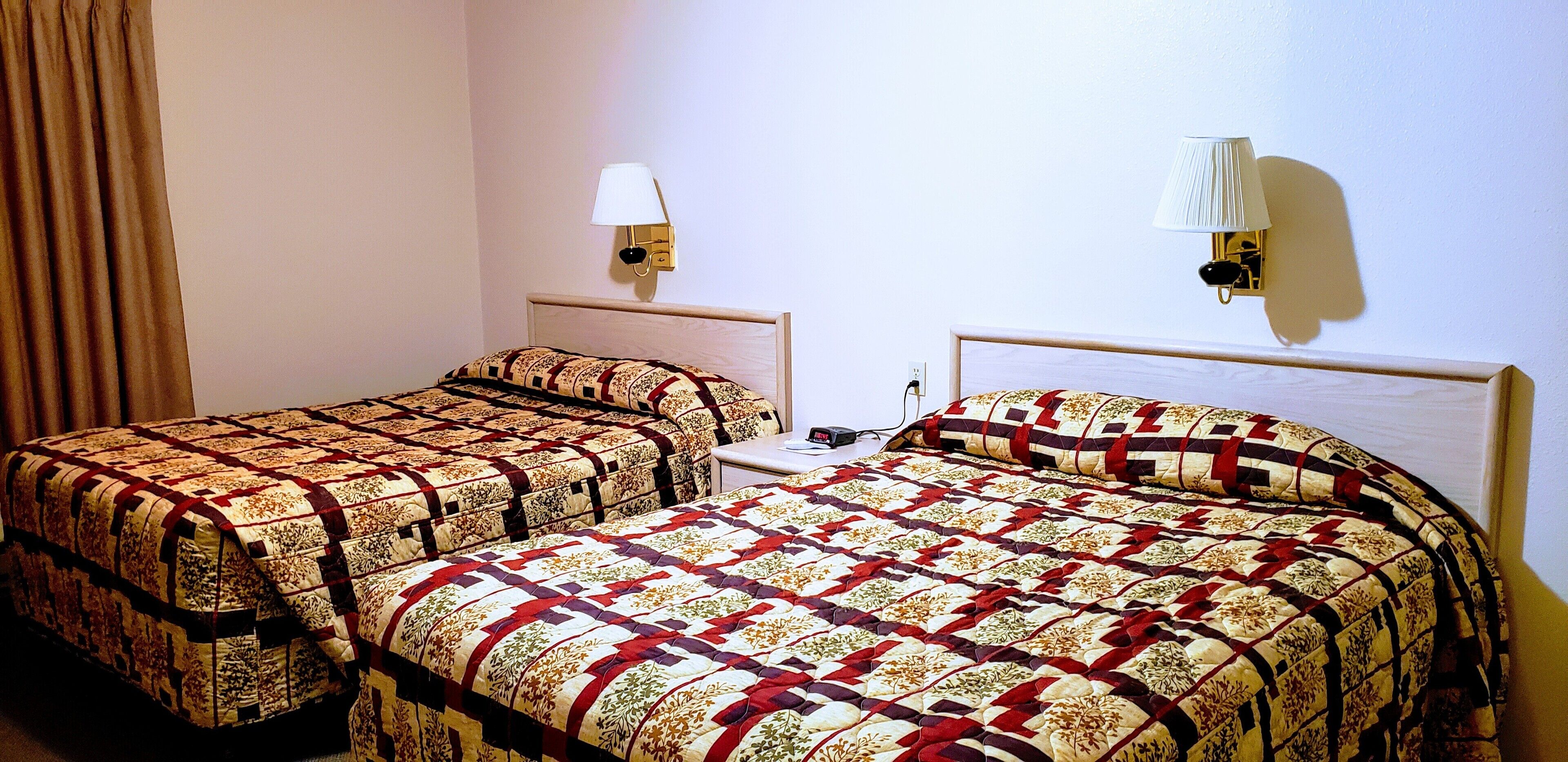 Una camera d'albergo con due letti, ognuno con una coperta a motivi e un comodino, e due lampade a parete.