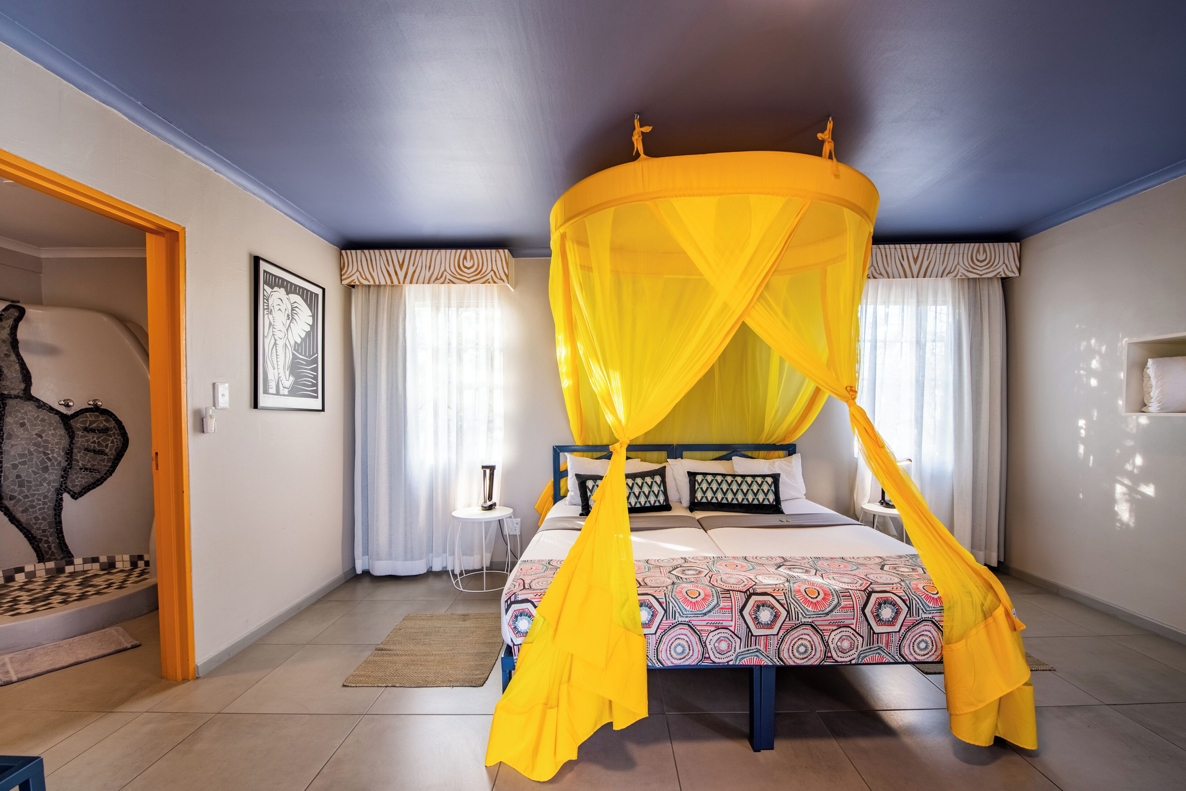 A bedroom with a bed under a yellow canopy, a nightstand, a lamp, and a wall art piece.
