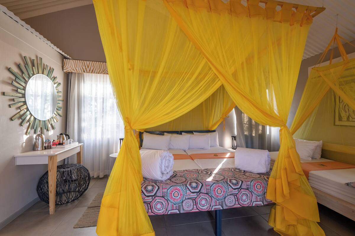 Quarto com cama sob um dossel amarelo, mesa de cabeceira com espelho e colcha estampada.