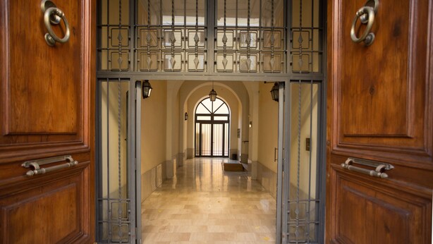 A grand entrance with a wooden door and metal gate, leading to a spacious hallway with a marble floor and an arched window at the end. - Romerooms (Rome)