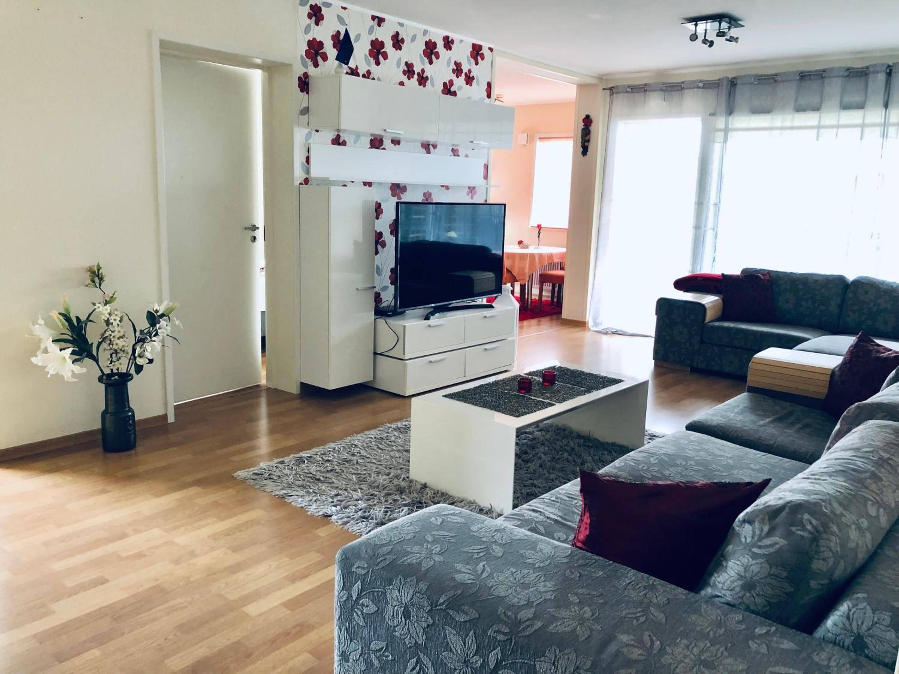 Uma sala de estar com sofá, televisão de tela plana, mesa de centro e um vaso decorativo.