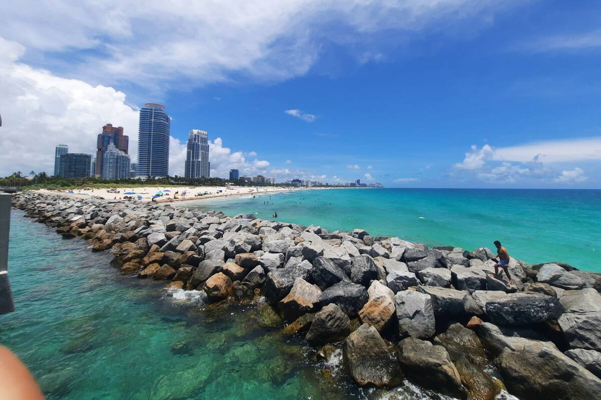 A coastal cityscape with a rocky shoreline, clear turquoise waters, and high-rise buildings in the background.