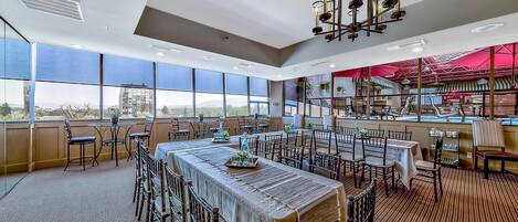 A spacious dining area with high ceilings and large windows offering a city view. Tables are set with chairs, and a chandelier hangs from the ceiling.