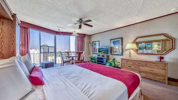 Habitación de hotel con una cama grande, ventilador de techo, escritorio con silla, cómoda y un gran ventanal con vista a la ciudad.
