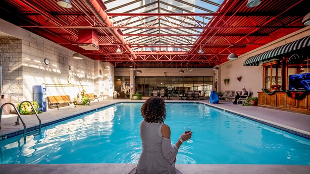 A pool area with a person sitting by the pool, a bar, and a view of the outside. - Plaza Resort Club (Reno)