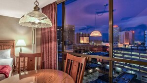 A hotel room with a large window offering a city view, a round wooden table, a chair, a bed with a wooden headboard, and a hanging lamp. - Plaza Resort Club (Reno)