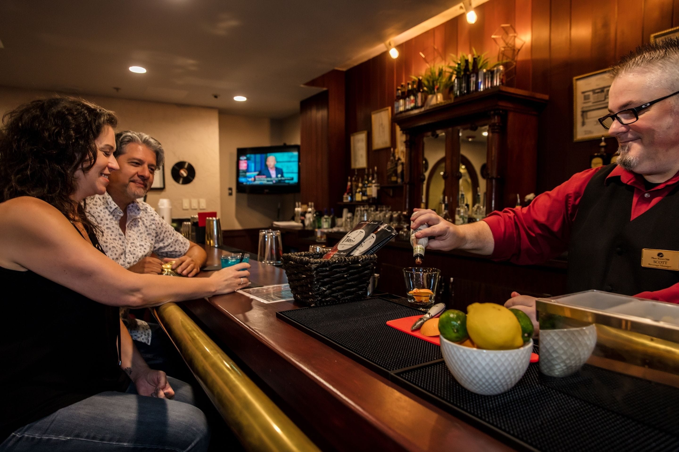 Een bar met een bartender, een televisie en een toonbank met verschillende flessen en een schaal fruit.