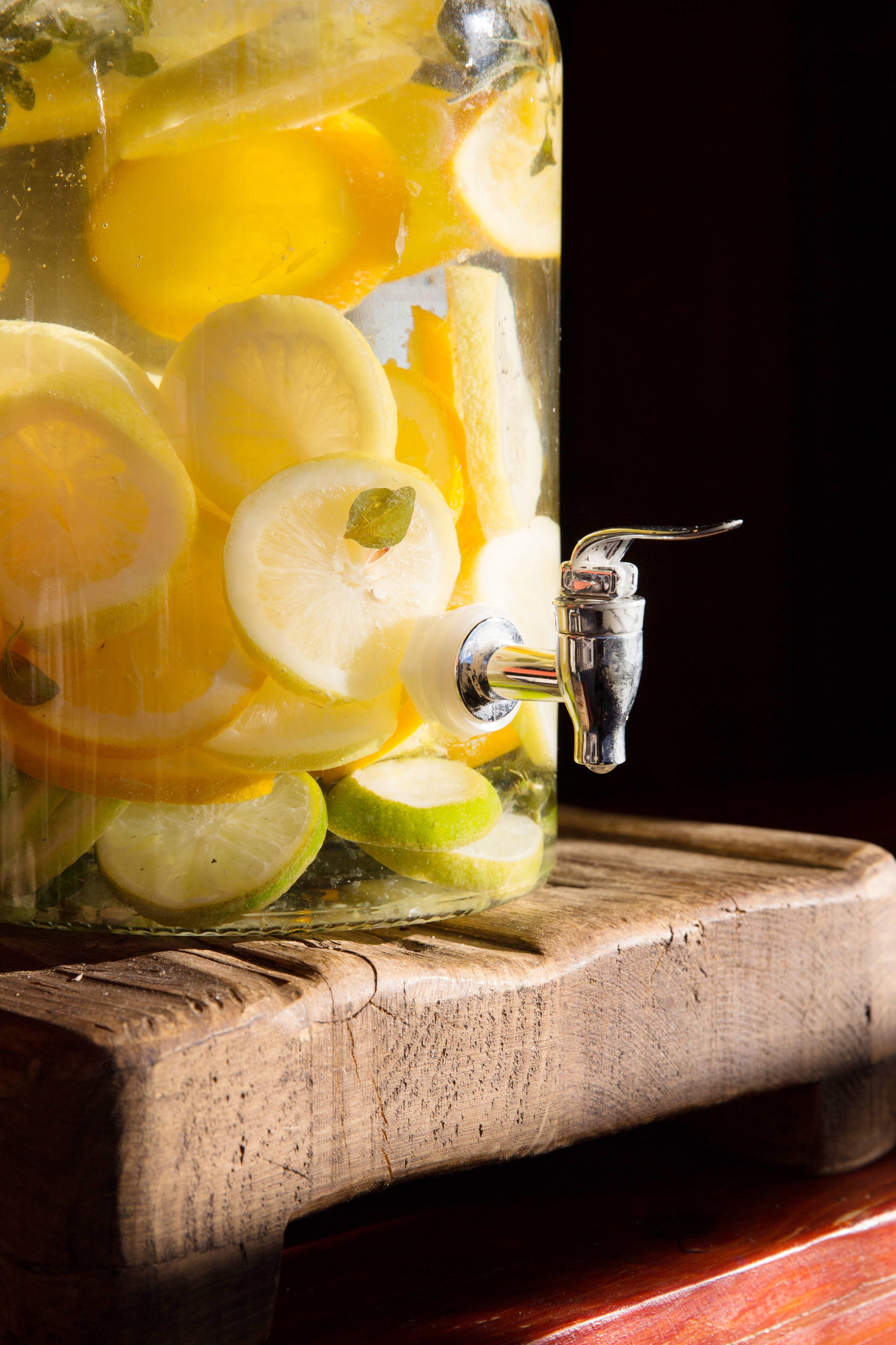 A clear glass container with a metal tap, filled with slices of citrus fruits and herbs, placed on a wooden surface.