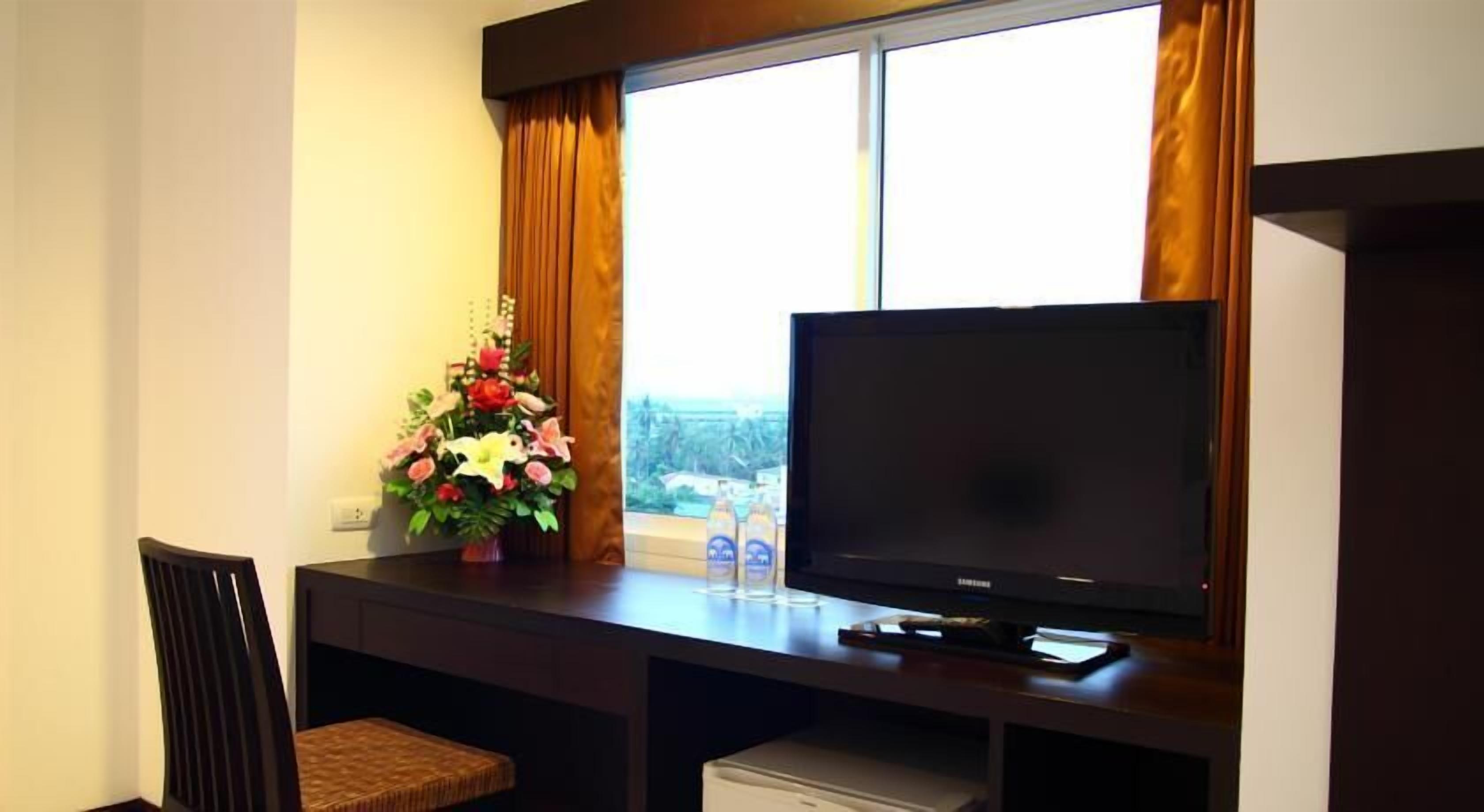 A hotel room with a TV on a wooden desk, a chair, a small table with a flower arrangement, and a window with curtains.
