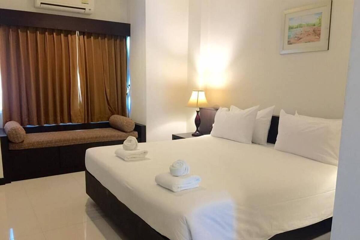 Una habitación de hotel con una cama grande, un banco con cojines, una lámpara de noche y un cuadro en la pared.