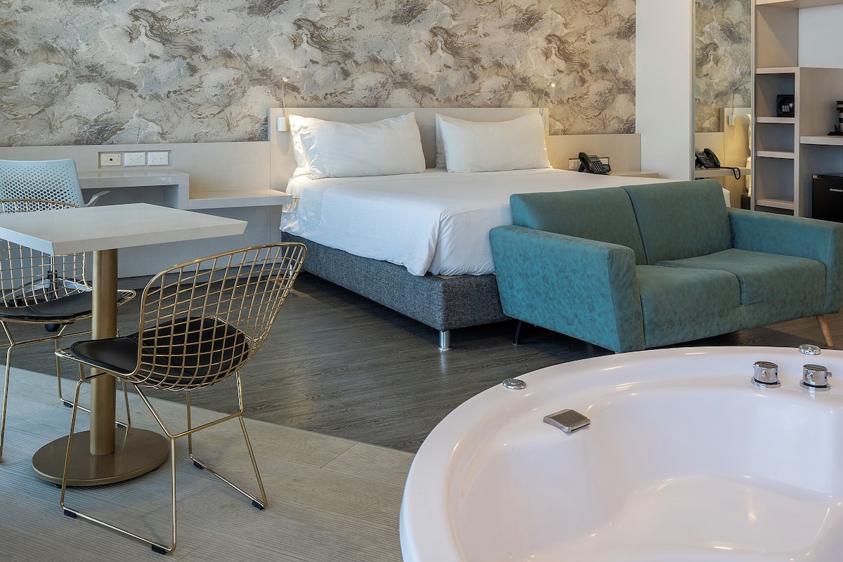 Una habitación de hotel moderna con cama, zona de estar con mesa y sillas, una bañera grande y una pared con diseño de mármol.