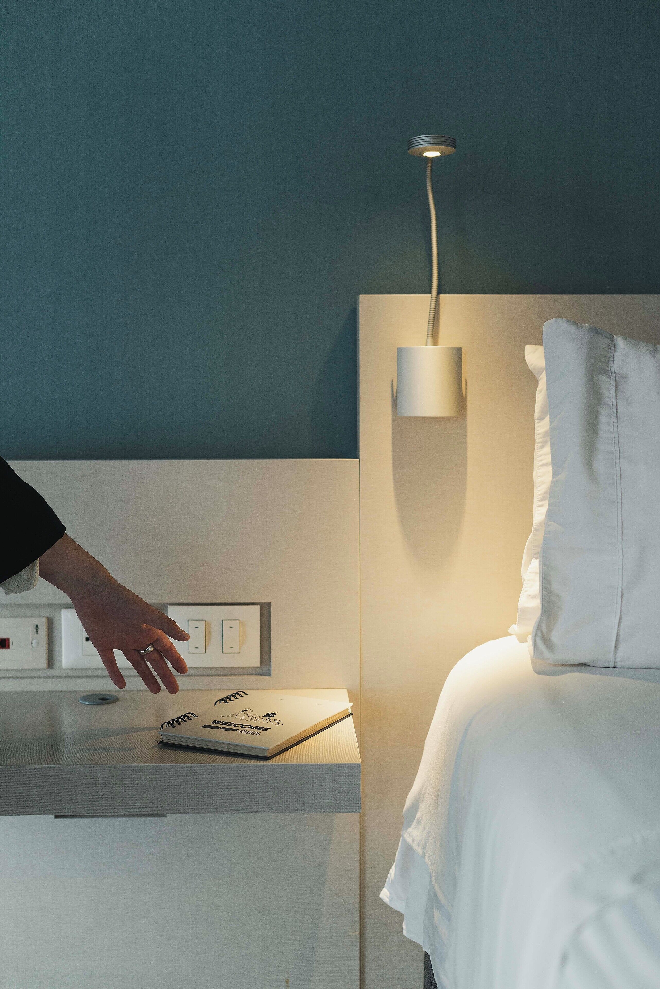 Una habitación de hotel moderna con mesita de noche, lámpara de pared y cama con ropa blanca.