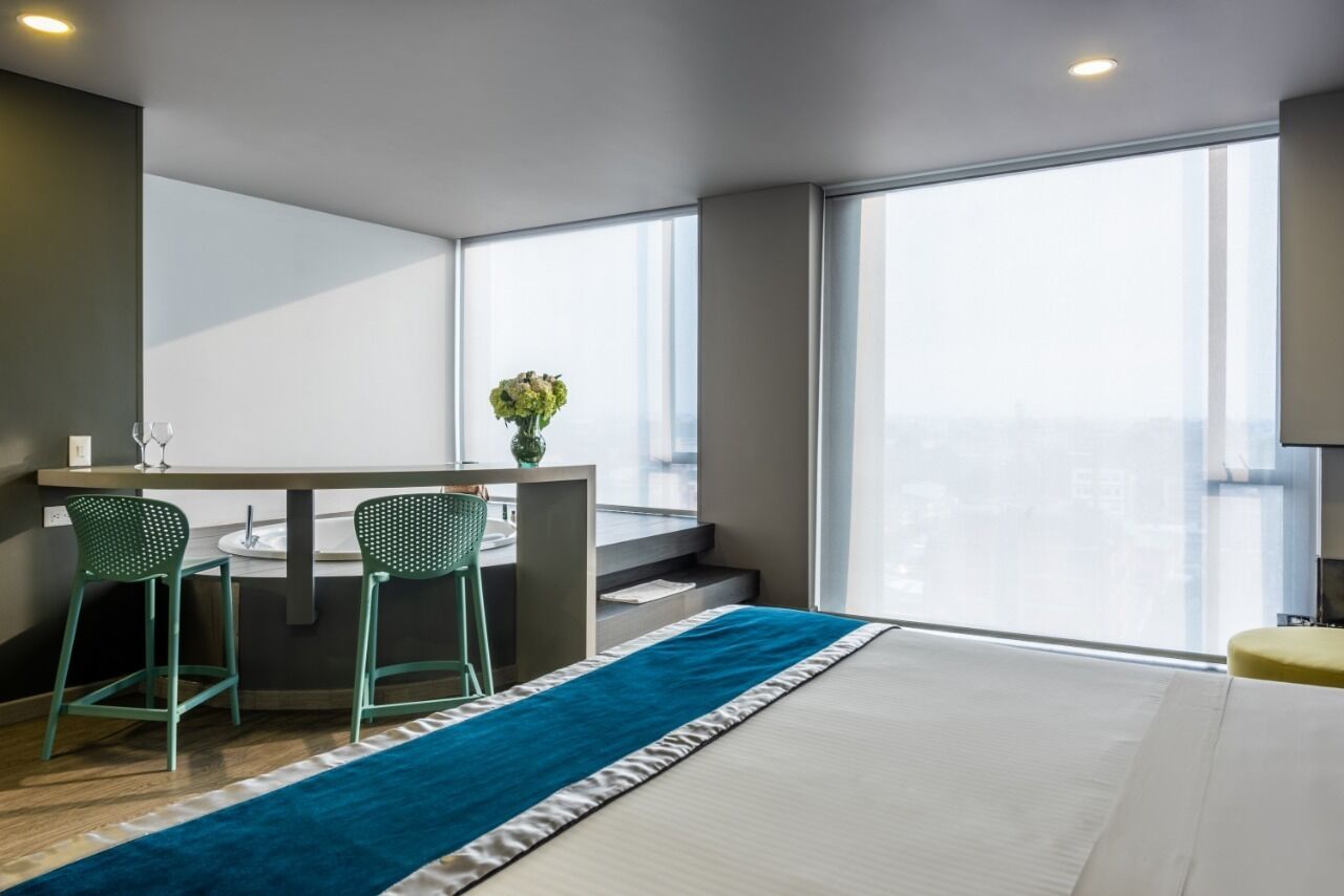 Een moderne hotelkamer met een groot raam, een klein kitchenette, twee groene stoelen en een blauw vloerkleed.