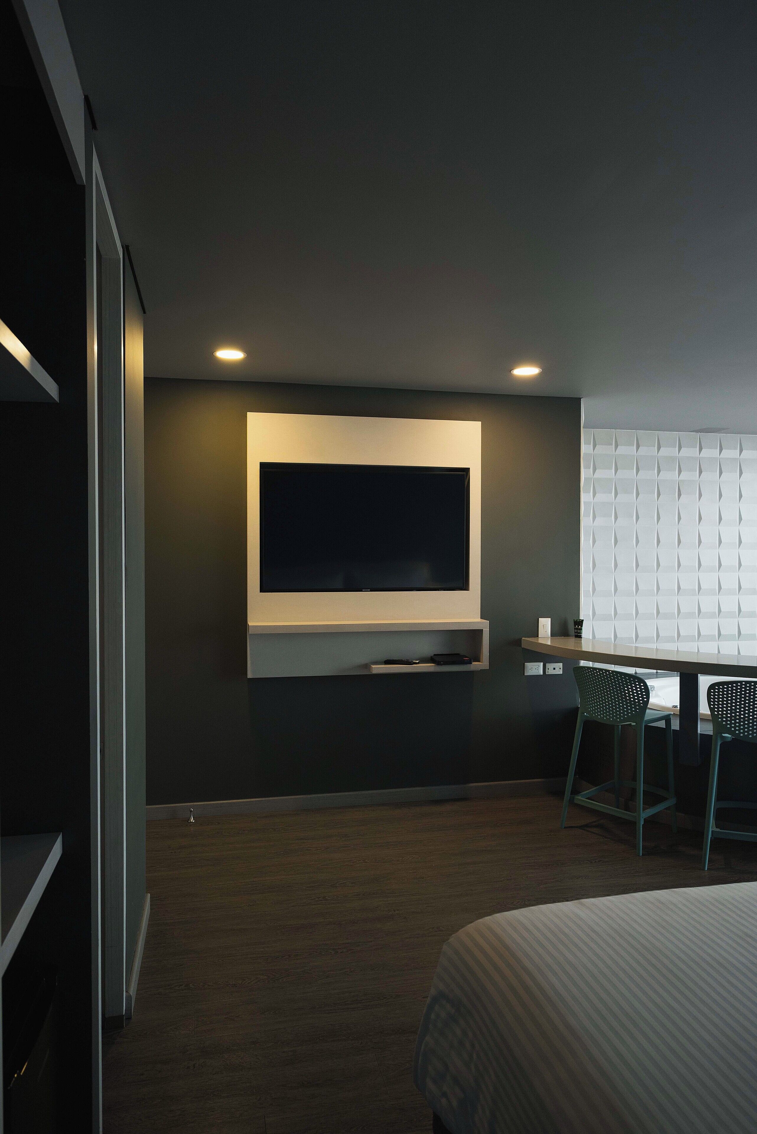 A modern hotel room with a large flat-screen TV mounted on the wall, a desk with a chair, and a bed with a striped bedspread.