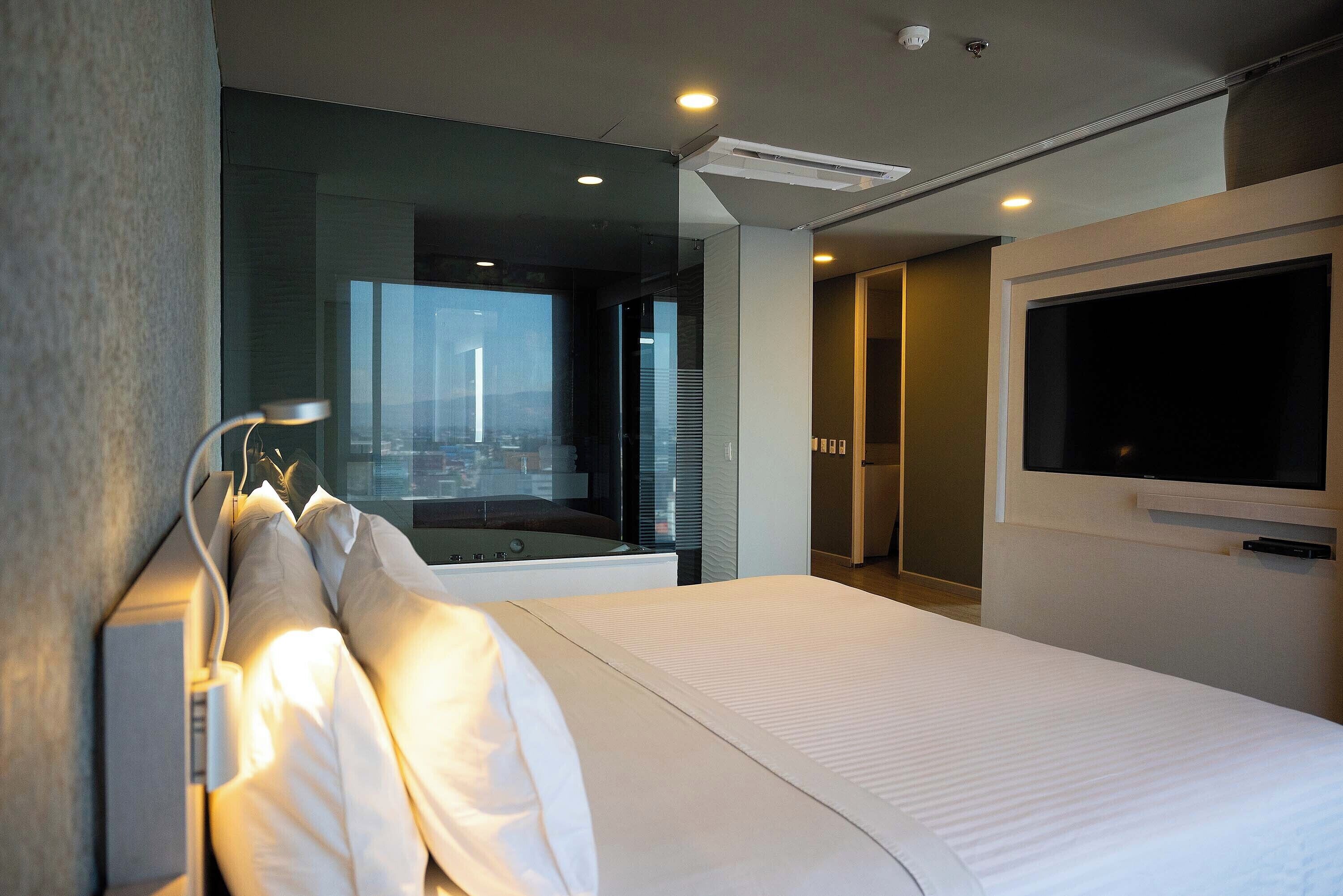 Habitación de hotel moderna con una cama grande, un televisor montado en la pared y vistas a la ciudad a través de la ventana.
