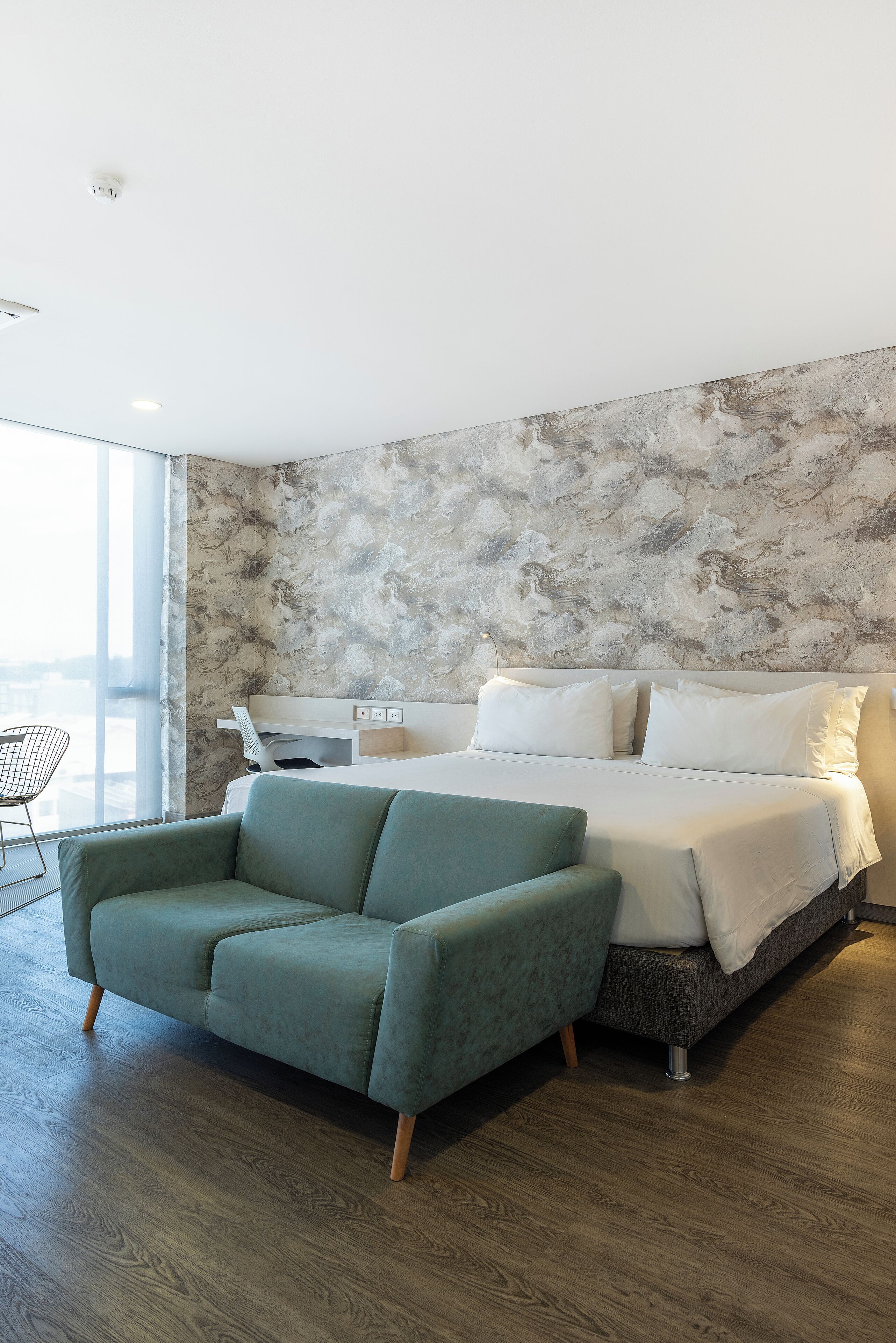 A modern bedroom with a large bed, a green sofa, and a city view through the window.
