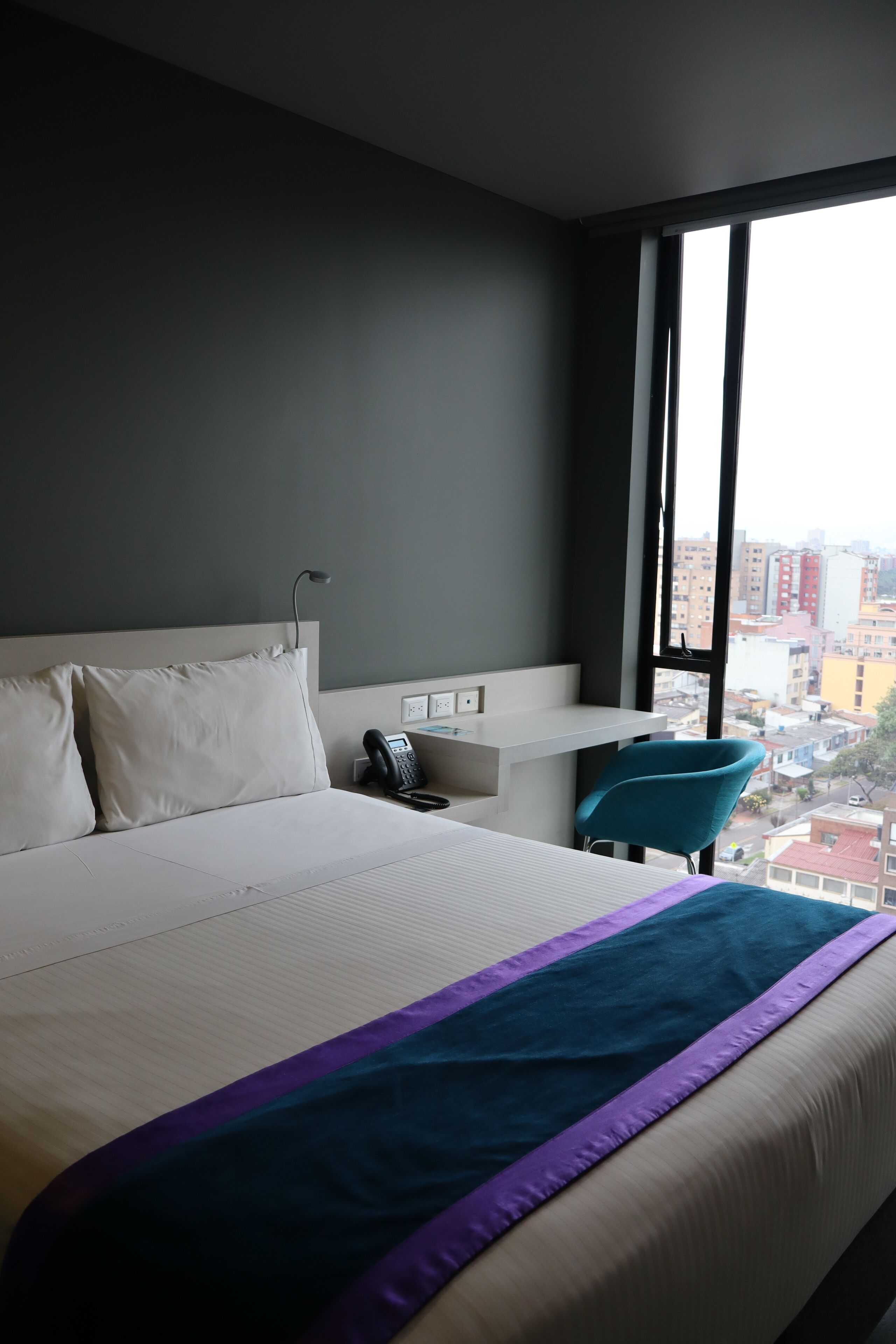 A hotel room with a large bed, a desk, a chair, and a view of the city through a large window.