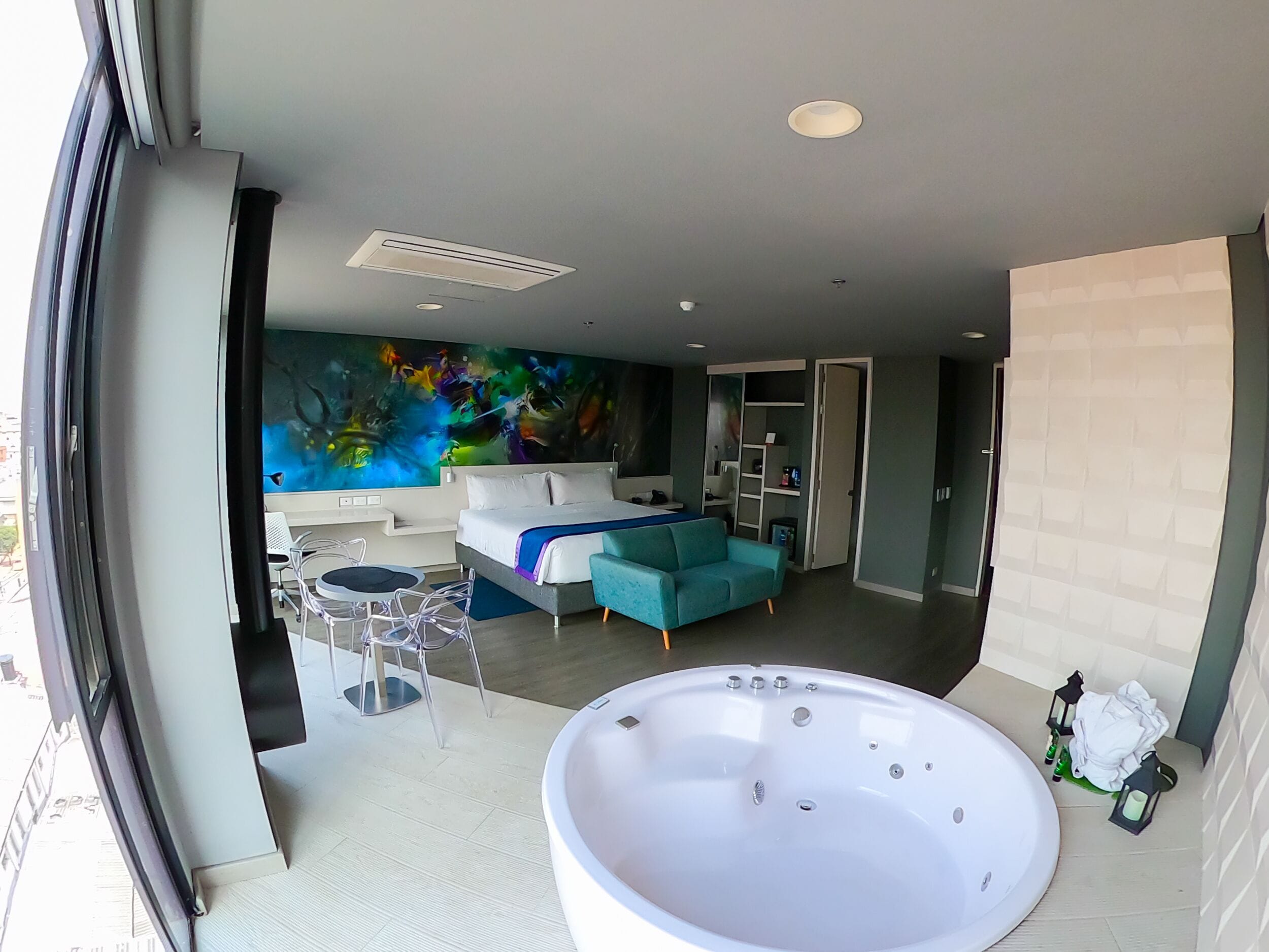 A modern hotel room with a large bed, a sitting area with a sofa and chairs, a large bathtub, and a vibrant abstract mural on the wall.