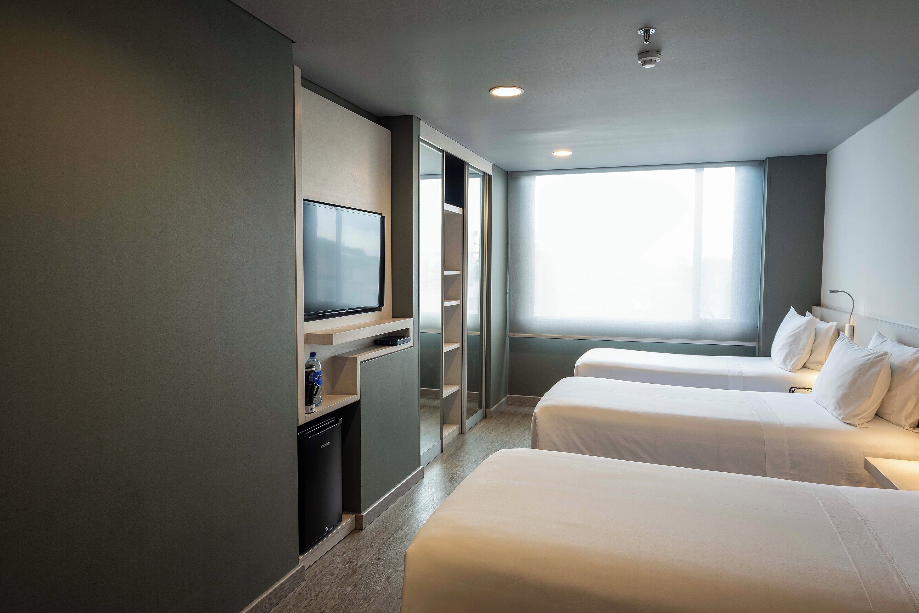 A hotel room with two beds, a TV, a minibar, and a large window providing natural light.