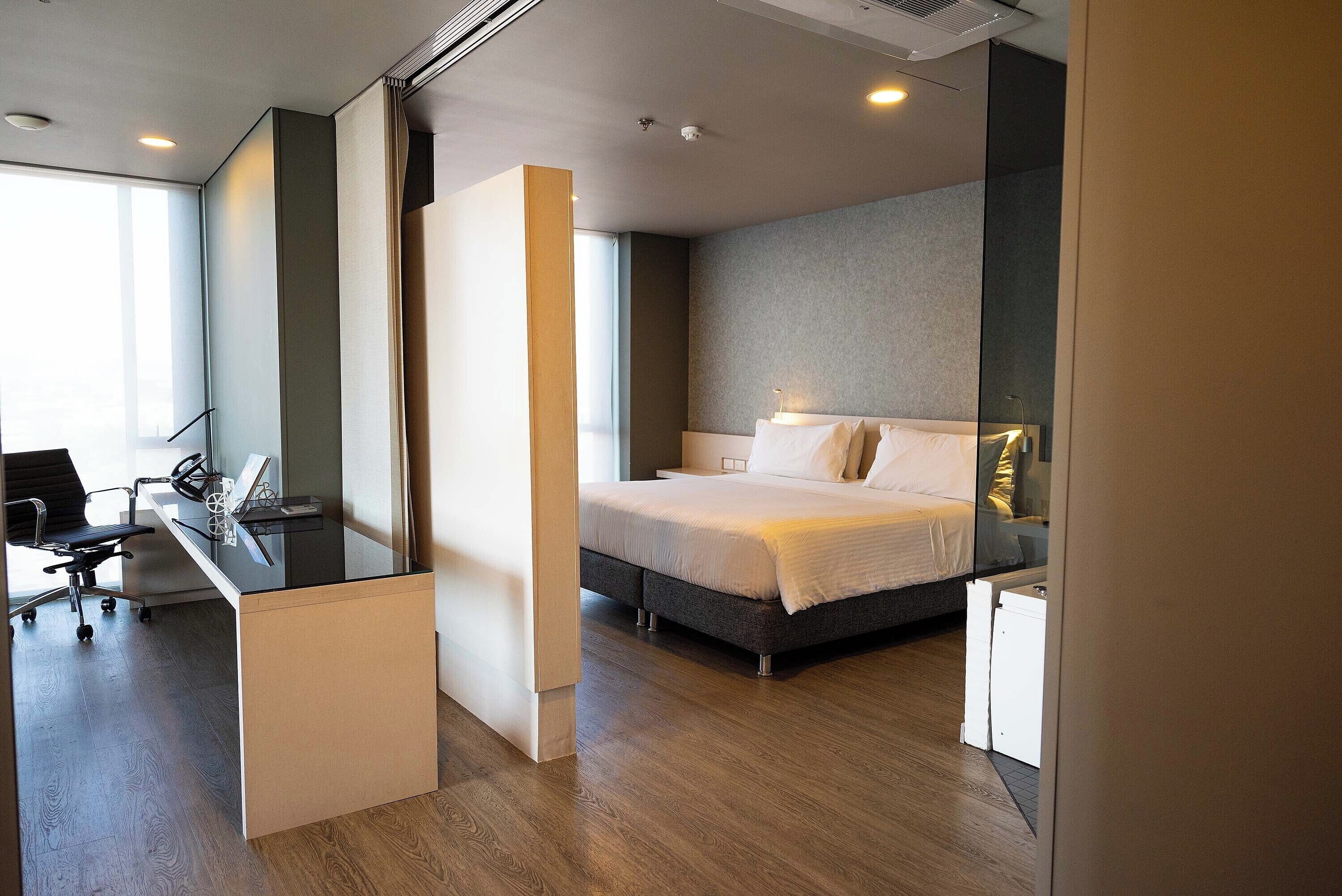 A modern hotel room with a bed, desk, and chair, featuring a large window and a view of the city.