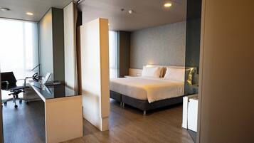A modern hotel room with a bed, desk, and chair, featuring a large window and a view of the city.
