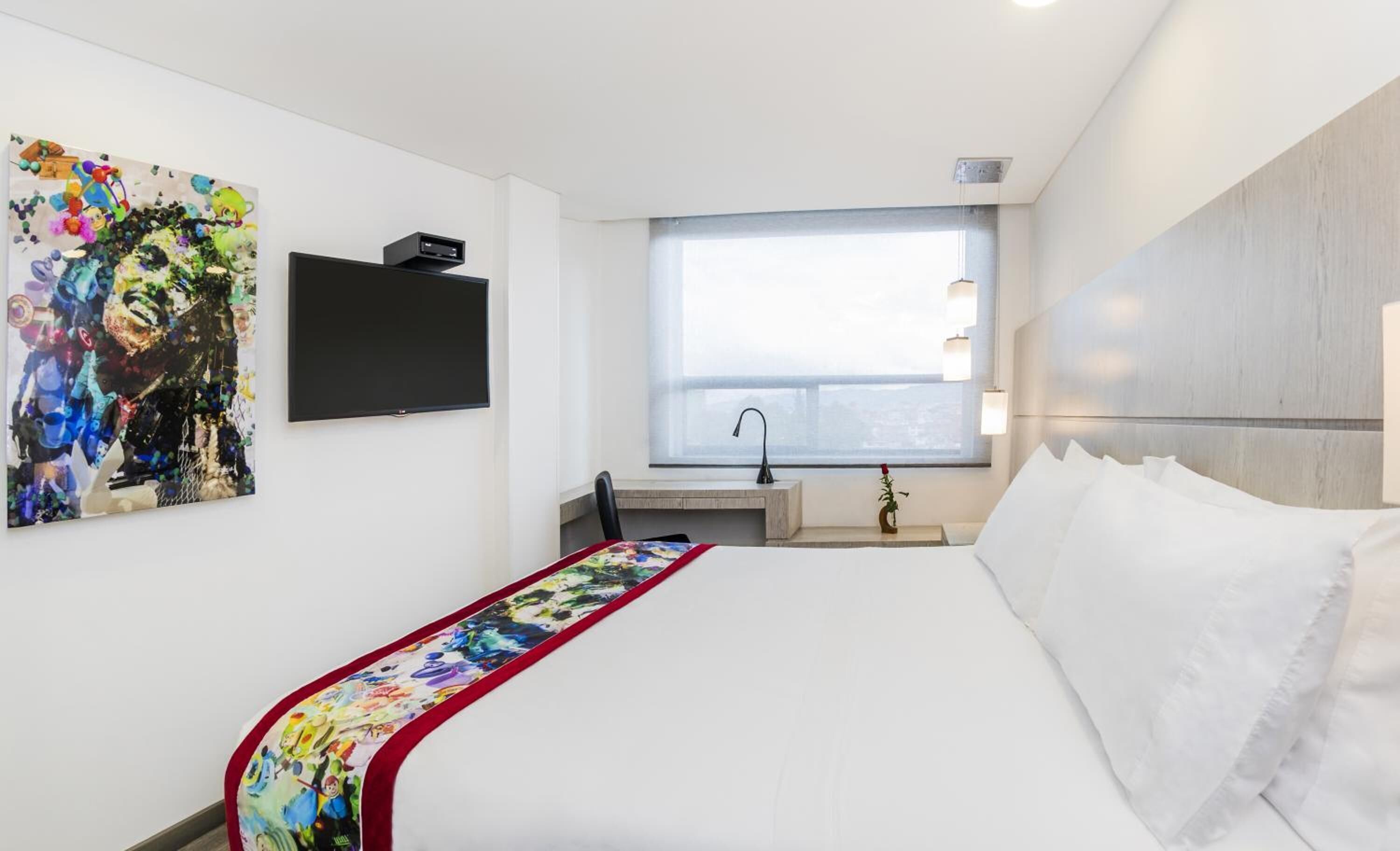 A hotel room with a bed, a desk, a window, and a TV on the wall.