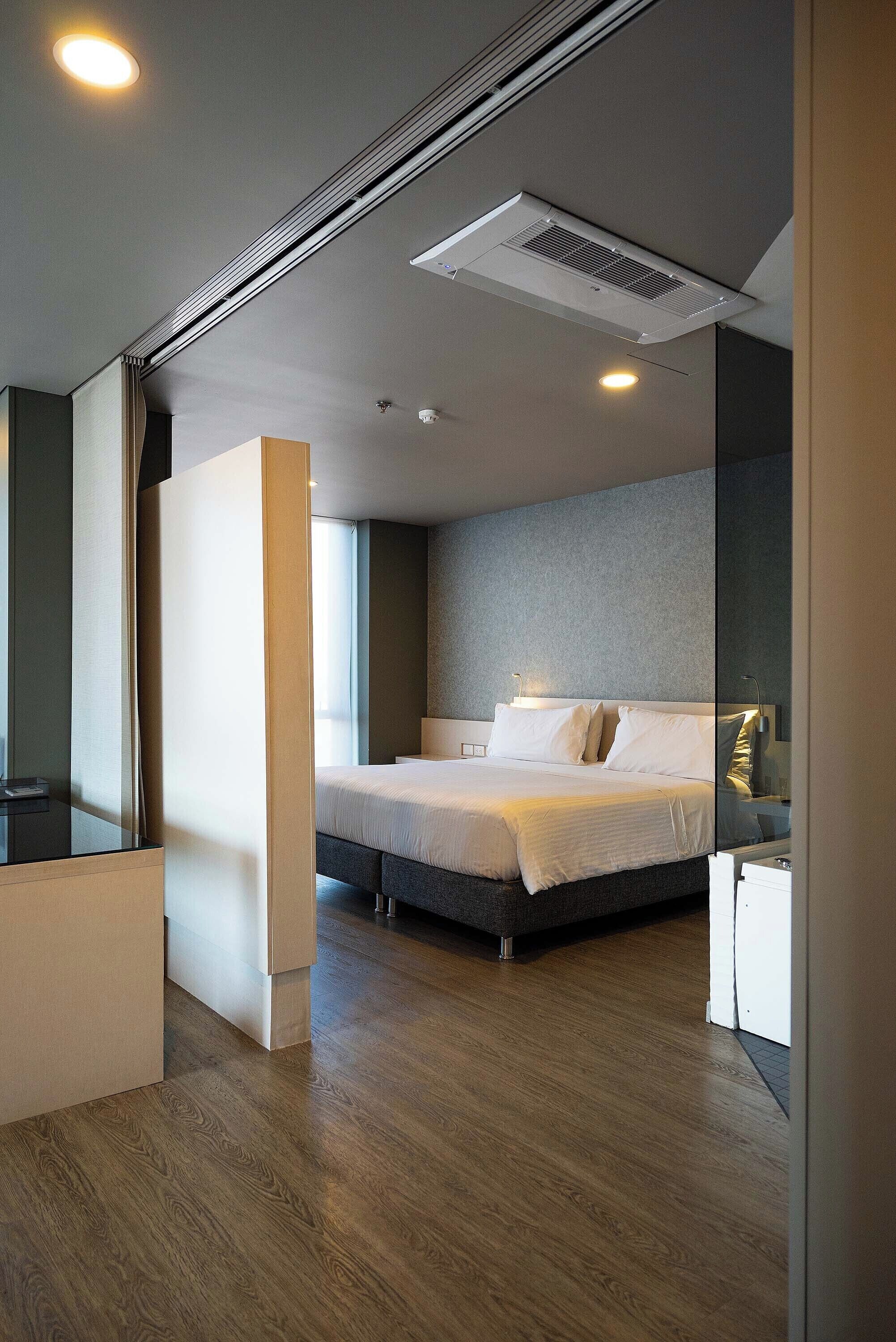 Una habitación de hotel moderna con una cama grande, una mesita de noche, un espejo y una rejilla de ventilación en el techo.