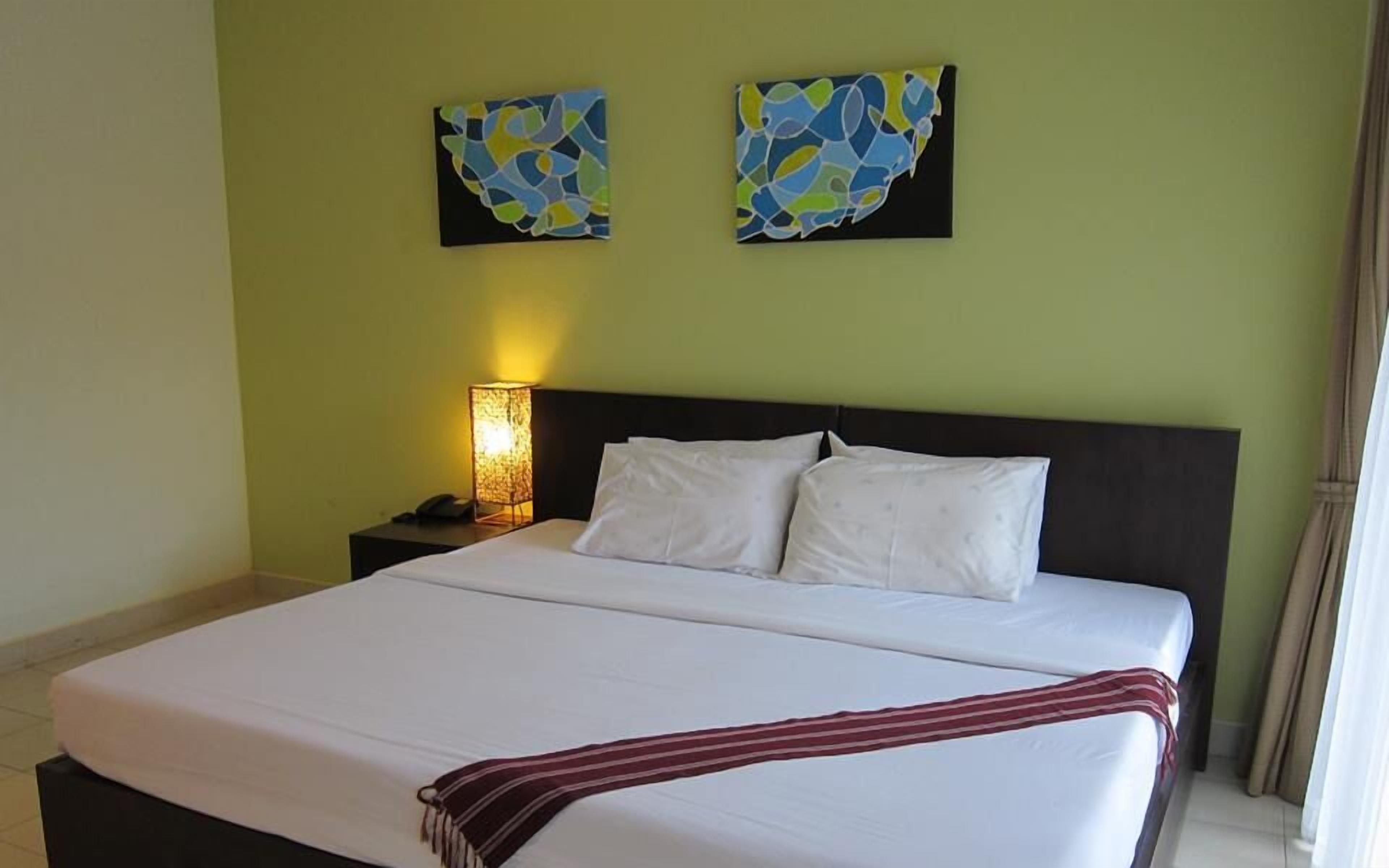 A hotel room with a large bed, white bedding, a striped throw blanket, a nightstand with a lamp, and abstract wall art.