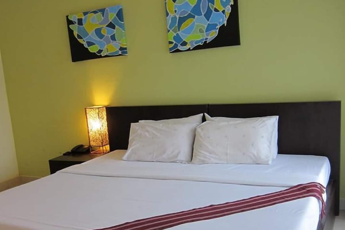 Une chambre d’hôtel avec un grand lit, deux oreillers, un plaid à rayures, une table de chevet avec une lampe et deux tableaux abstraits accrochés au mur.