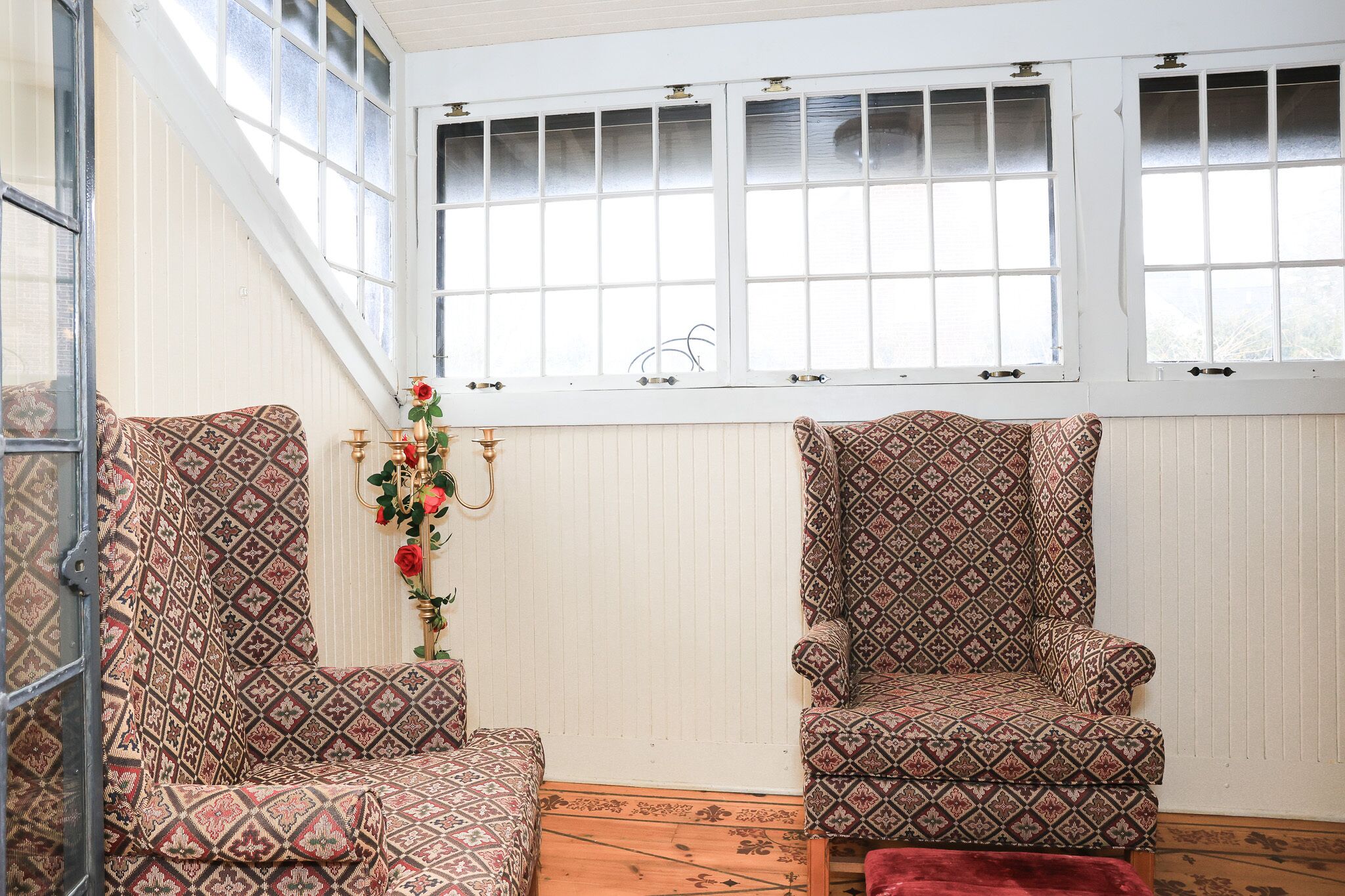Una habitación con dos sillones estampados, suelo de madera, una ventana con persianas y un jarrón decorativo con flores.