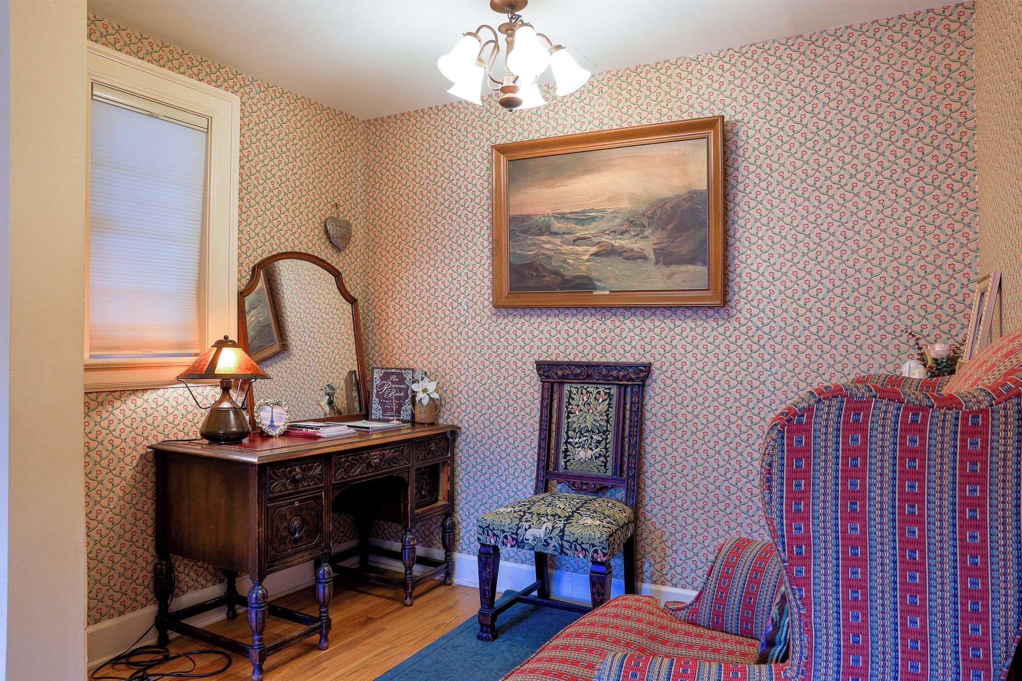 A room with patterned wallpaper, a wooden desk with a lamp, a chair, a bed with a patterned bedspread, and a framed painting on the wall.