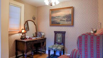 A room with patterned wallpaper, a wooden desk with a lamp, a chair, a bed with a patterned bedspread, and a framed painting on the wall.