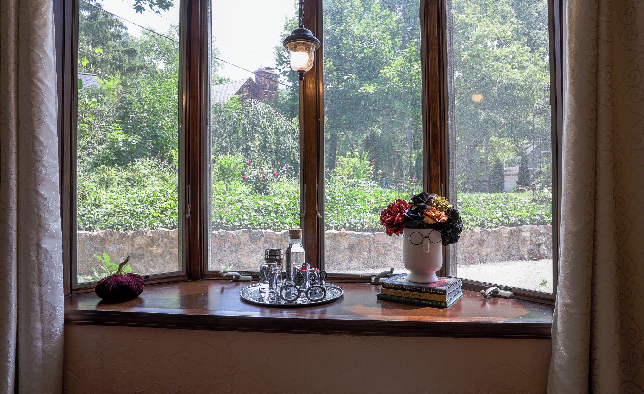 Janela com vista para vegetação, uma lâmpada, um abóbora e livros no parapeito.