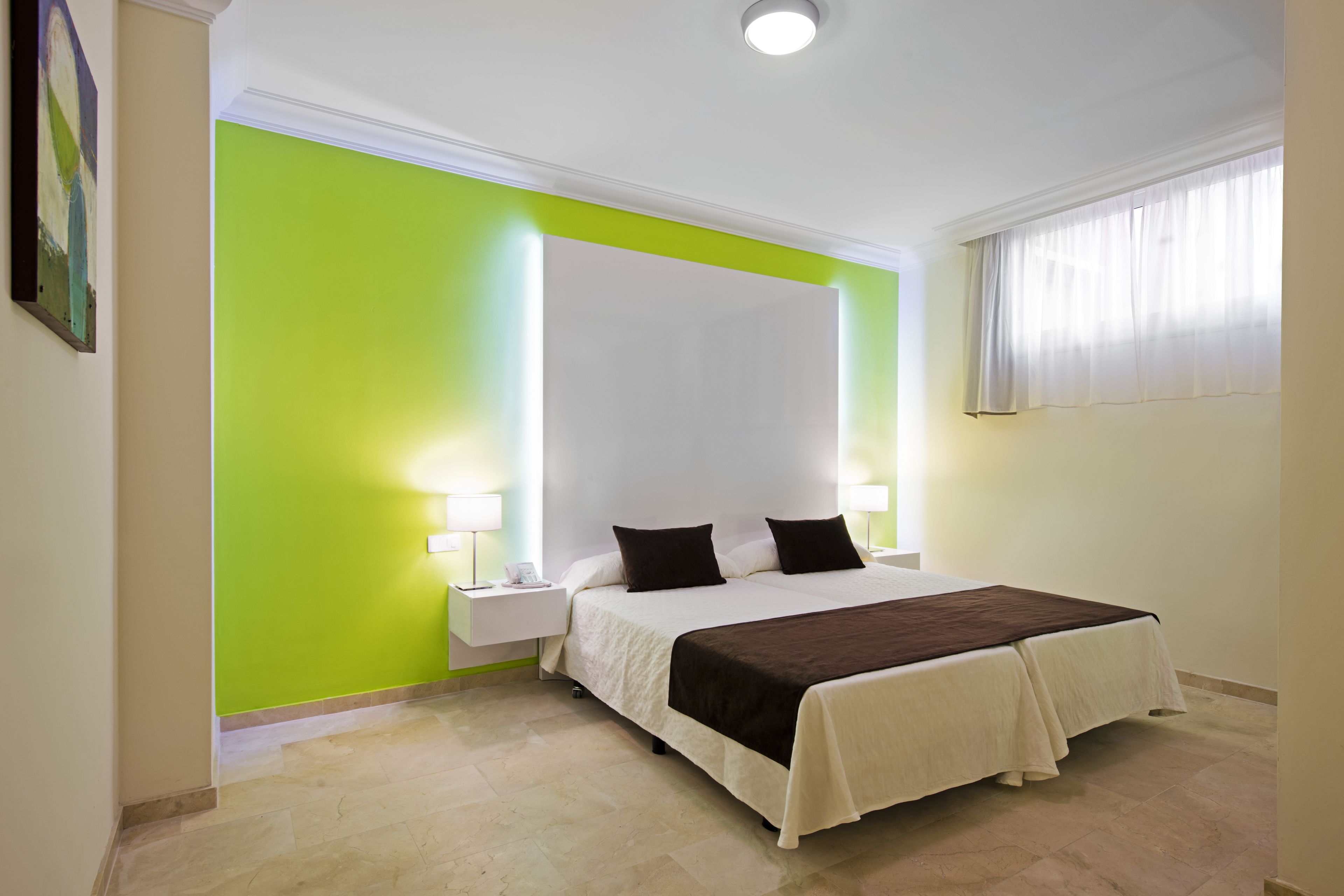 Une chambre d’hôtel avec un grand lit, deux tables de chevet avec des lampes, un mur d’accent vert et une fenêtre avec des rideaux.