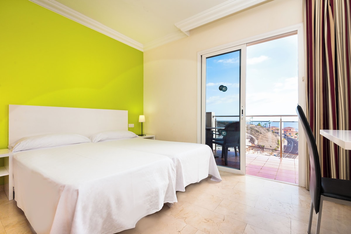 A hotel room with a large bed, a bright green wall, and a balcony with a view of the city.