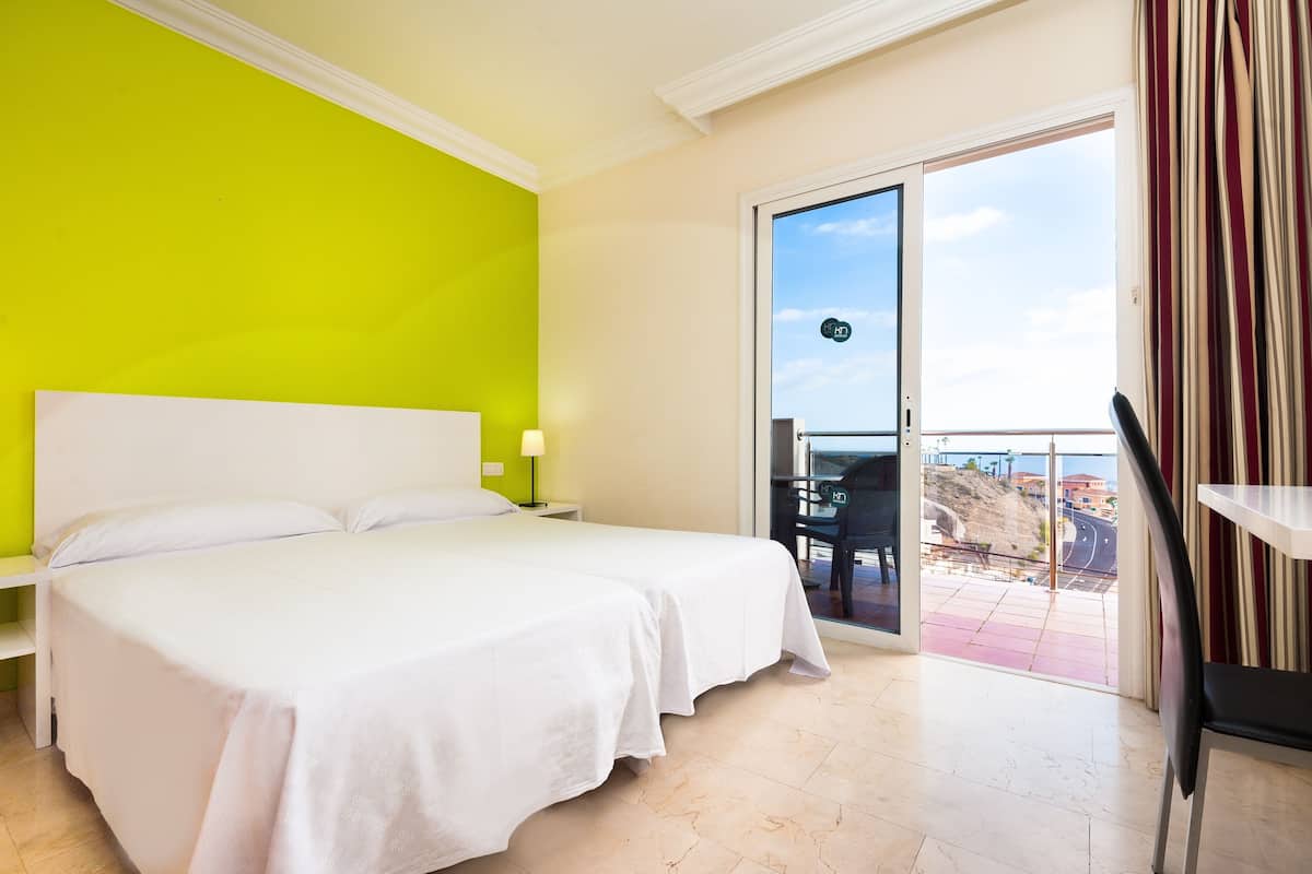 A hotel room with a large bed, a bright green wall, a balcony with a view, and a chair.