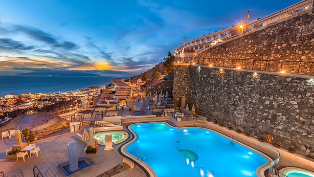 A swimming pool with a stone wall and a view of the city at dusk. - Kn Aparthotel Panoramica Heights (Adeje)