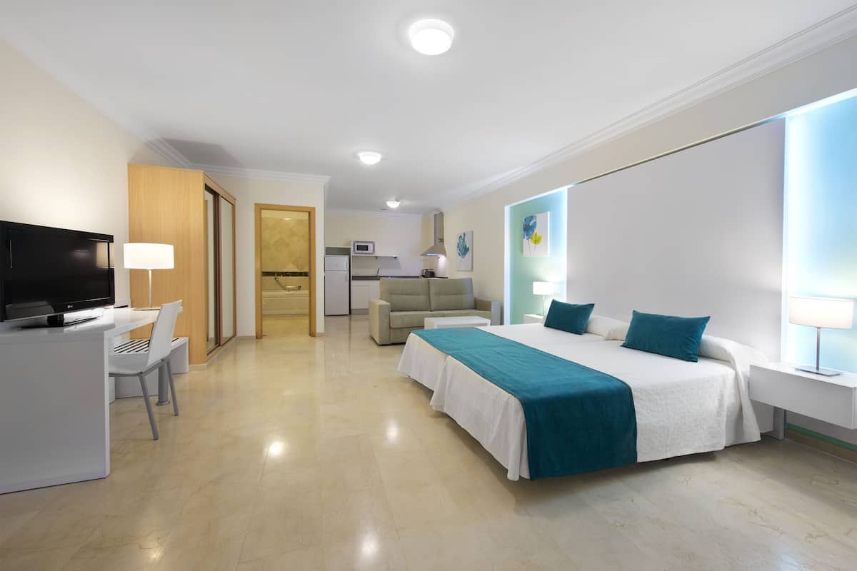A modern hotel room with a bed, desk, chair, television, and kitchenette.