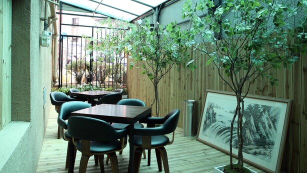 Eine überdachte Terrasse mit Holztischen und -stühlen, einem gerahmten Gemälde und einem kleinen Baum.