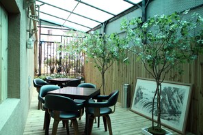 Eine überdachte Terrasse mit Holztischen und -stühlen, einem gerahmten Gemälde und einem kleinen Baum.