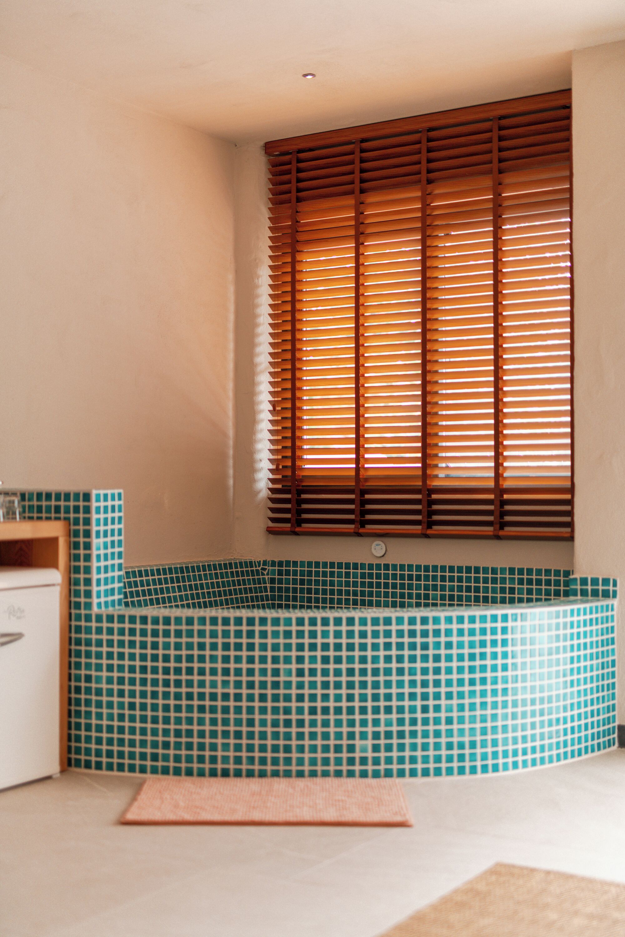 A bathroom with a bathtub, a window with blinds, and a tiled wall.