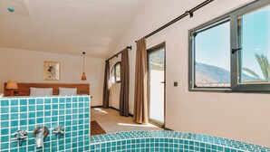 A hotel room with a large window, a bed with a headboard, a bathroom with a tiled wall, and a view of the outdoors. - Ünsal Hotel (Fethiye)