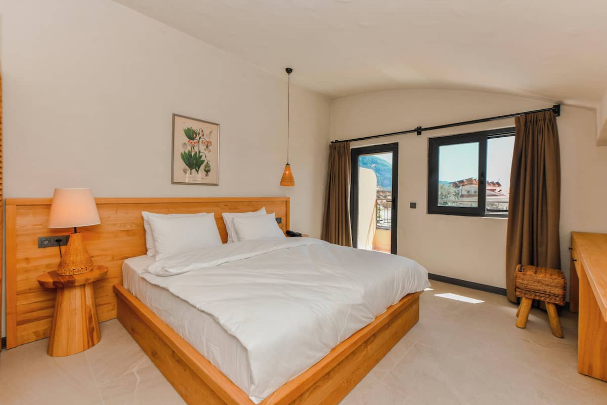 Un dormitorio con una cama de madera, una mesita de noche con lámpara, un grabado botánico enmarcado en la pared y vistas a través de una ventana con cortinas.