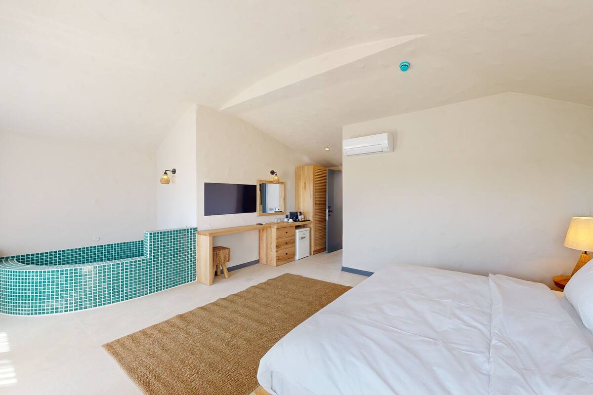 Habitación de hotel con cama, escritorio, televisor, kitchenette pequeña y baño con bañera azulejada.