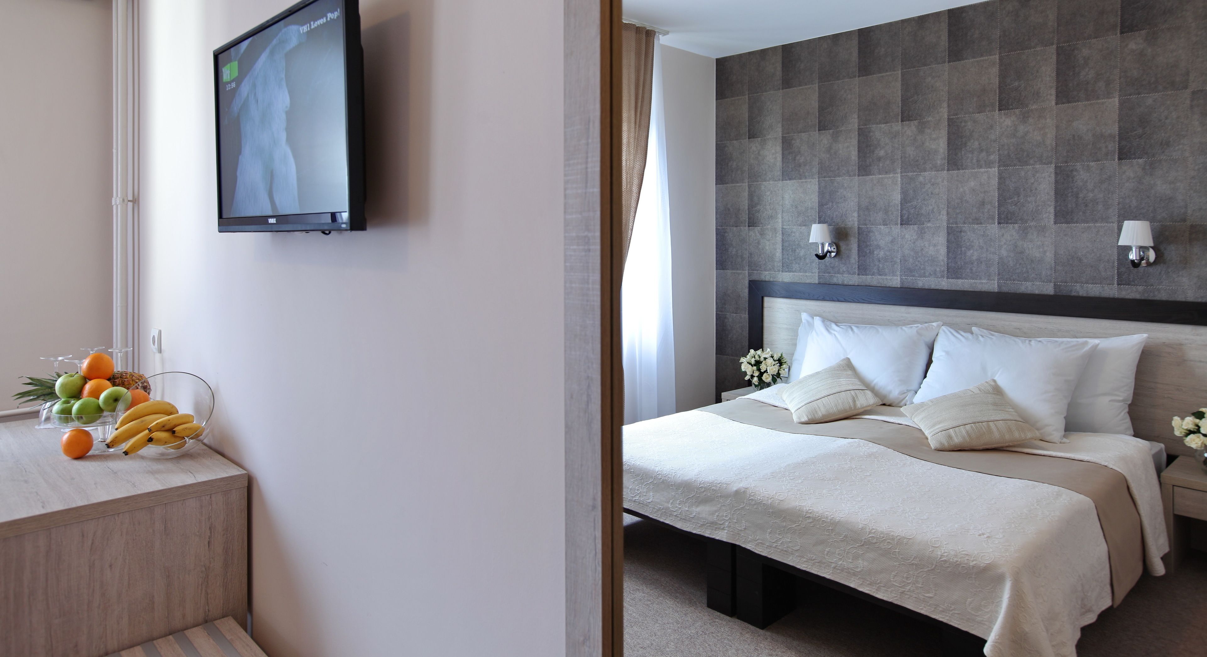 Quarto de hotel com uma cama, abajures, uma TV na parede e uma fruteira no criado-mudo.