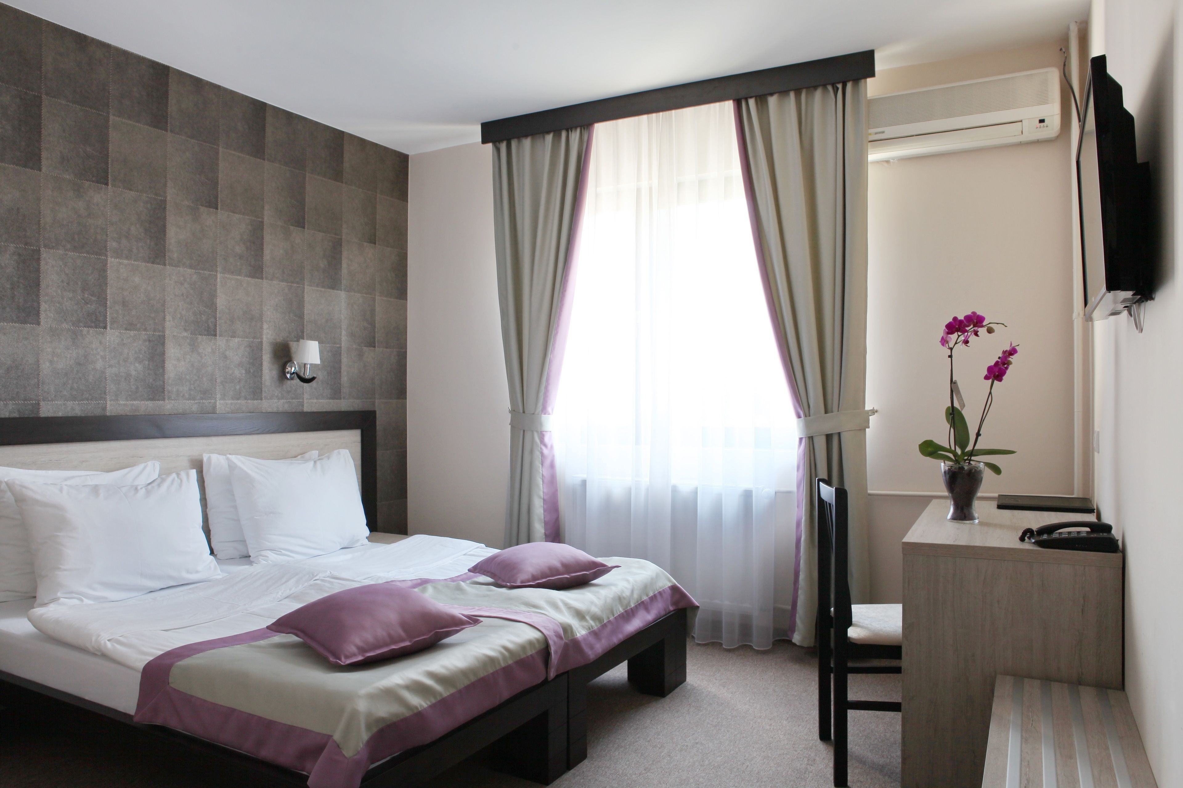 Habitación de hotel con cama, mesita de noche, una silla, televisor y ventana con cortinas.