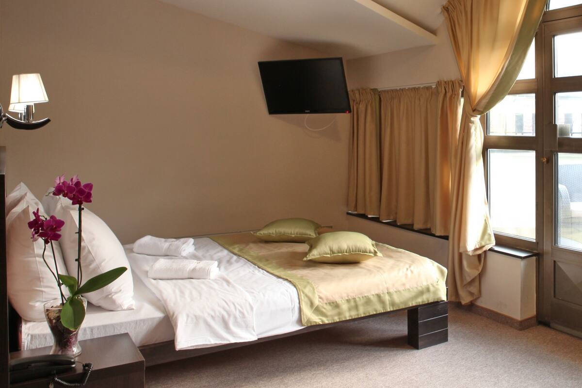 A hotel room with a bed, a bedside table with a lamp, a TV mounted on the wall, and a window with curtains.