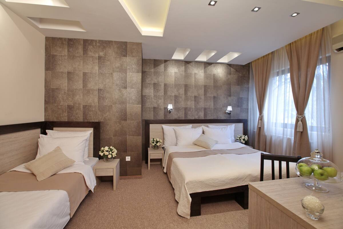 A hotel room with two beds, a wooden headboard, a small table with a glass bowl of apples, and a wall-mounted lamp.